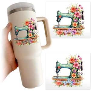 May include: A cream-colored insulated tumbler with a handle, featuring a floral design and a teal sewing machine. The design includes the name "ashley" in cursive script, surrounded by pink, orange, and purple flowers.