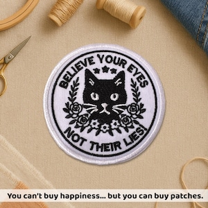 Peut inclure: Un écusson rond blanc avec un chat noir brodé et le texte "BELIEVE YOUR EYES NOT THEIR LIES". Le chat a des yeux et des moustaches blancs, entouré de motifs floraux et de feuilles. L'écusson est sur une surface beige.