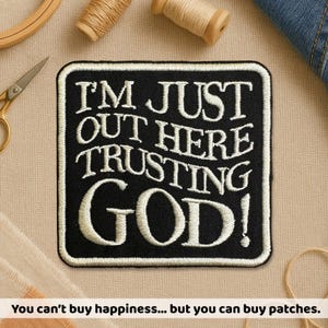 May include: Black embroidered patch with the text "I'M JUST OUT HERE TRUSTING GOD!" in white. The patch has a black background and a white border. The patch is surrounded by sewing supplies.