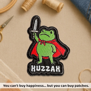 May include: Embroidered patch featuring a green frog wearing a red cape and holding a sword. The patch has a black border and the word "Huzzah" at the bottom. The image also includes the text: "You can't buy happiness... but you can buy patches."