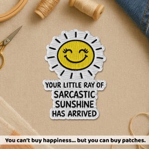 May include: Embroidered patch featuring a smiling sun with the text "YOUR LITTLE RAY OF SARCASTIC SUNSHINE HAS ARRIVED." The sun is yellow with a white outline and black details. The patch is on a beige background with sewing supplies.