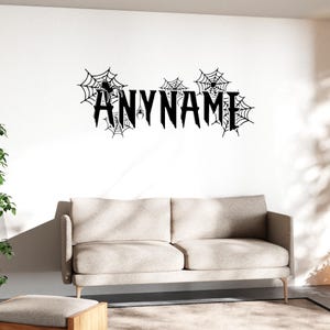 Peut inclure: Décoration murale noire avec le mot "ANYNAME" en police gothique, avec des toiles d'araignées et des araignées. L'œuvre est au-dessus d'un canapé beige.