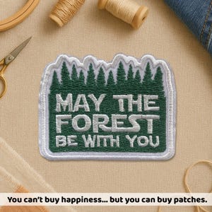 May include: Embroidered patch with the text "MAY THE FOREST BE WITH YOU" in white on a green and white background. The patch features a forest design at the top. Sewing supplies are in the background.