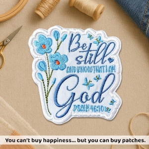 May include: Embroidered patch featuring the words "Be still and know that I am God" in blue script, with blue flowers and butterflies. The patch has a white background and a blue border. The text "Psalm 46:10" is also included.