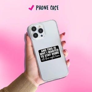 May include: Clear phone case with a black and white sticker that reads "AND THIS IS WHY I WANTED TO STAY HOME all this right here." The phone case is held in a hand against a pink background. The text "PHONE CASE" is at the top.
