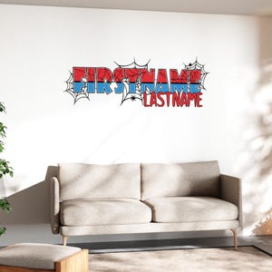 Peut inclure: Un autocollant mural avec le texte "FIRSTNAME LASTNAME" en rouge et bleu, avec des toiles d'araignées et des araignées. Le texte est sur un mur blanc au-dessus d'un canapé et d'un pouf beiges.