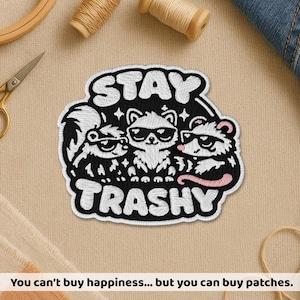 May include: Embroidered patch with the words "STAY TRASHY" in white lettering. The patch features three cartoon raccoons wearing sunglasses. The patch is black and white with a pink accent. The patch is on a tan fabric.