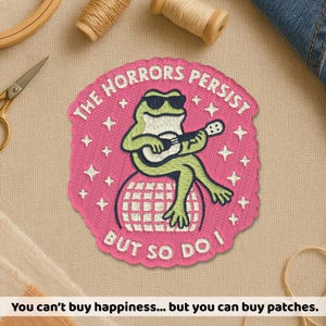 May include: Embroidered patch with a pink background and the text "THE HORRORS PERSIST BUT SO DO I". The patch features a green frog wearing sunglasses and playing a guitar while sitting on a disco ball. White stars are scattered around the frog.