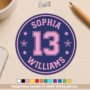 May include: A round sticker with a navy blue background features the name "SOPHIA" above the number "13" and the name "WILLIAMS" below. The text and number are in pink, with pink stars around the edge. The sticker also has the text "Happiness comes in small sticky pieces."