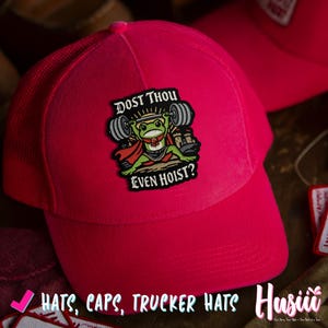 Puede incluir: Gorra de camionero rosa brillante con la parte trasera de malla. La parte delantera presenta un parche con una rana de dibujos animados levantando pesas, el texto dice "DOST THOU EVEN HOIST?" y las palabras "HATS, CAPS, TRUCKER HATS" est&aacute;n en la parte inferior.