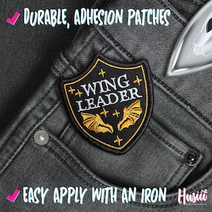 May include: A black and gold embroidered patch with the words "WING LEADER" and bat wings. The patch is on a pair of dark gray jeans. The text "DURABLE, ADHESION PATCHES" and "EASY APPLY WITH AN IRON" are also visible.