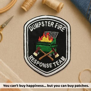 May include: Embroidered patch with a black shield shape, featuring a green dumpster on fire with crossed axes below. Text reads "DUMPSTER FIRE RESPONSE TEAM" in white. The bottom of the image reads "You can't buy happiness... but you can buy patches."