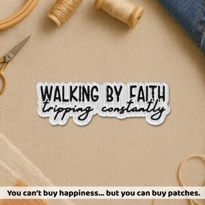 May include: Embroidered patch with the text "WALKING BY FAITH tripping constantly" in black on a white background. The patch is rectangular with a wavy border. Sewing supplies and denim are in the background.