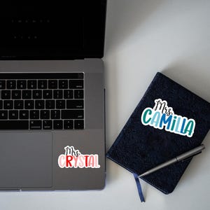 K&ouml;nnte beinhalten: Ein Laptop mit einem Aufkleber, auf dem "Mrs. Crystal" steht, auf dem Trackpad. Ein schwarzes Notizbuch mit einem Aufkleber, auf dem "Mrs. Camilla" steht, auf dem Einband.