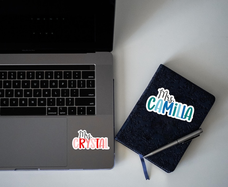 K&ouml;nnte beinhalten: Ein Laptop mit einem Aufkleber, auf dem "Mrs. Crystal" steht, auf dem Trackpad. Ein schwarzes Notizbuch mit einem Aufkleber, auf dem "Mrs. Camilla" steht, auf dem Einband.