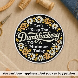 Op de afbeelding: Ronde geborduurde patch met de tekst "Let's Keep The Dumbfuckery To A Minimum Today" in witte letters op een zwarte achtergrond. De patch heeft een rand van witte en gele bloemen. Naaibenodigdheden omringen de patch.