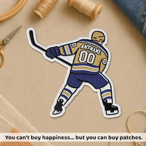 May include: Embroidered hockey player patch in blue, gold, and white. The patch features the name "ANYNAME" above the number "00" on the jersey. The patch is on a tan fabric surface with sewing supplies.
