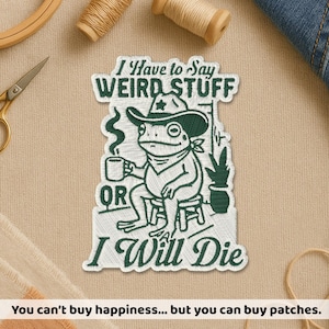 Peut inclure: Un écusson brodé blanc avec des coutures vert foncé représentant une grenouille portant un chapeau de cowboy et un bandana, assise sur un tabouret avec une tasse fumante. Le texte indique I Have to Say WEIRD STUFF OR I Will Die avec une plante.
