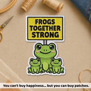 May include: Embroidered patch featuring three green frogs. The largest frog holds a sign that reads "Frogs Together Strong" in black text on a yellow background. The patch has a white border and is likely for clothing or accessories.