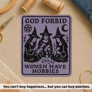 May include: A lavender patch with the text "GOD FORBID WOMEN HAVE HOBBIES" and an illustration of three witches. The patch has a pentagram and crescent moon design. The bottom text reads "You can't buy happiness... but you can buy patches."