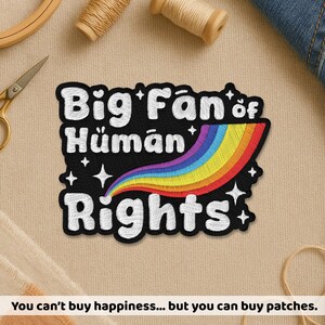 May include: Embroidered patch with the text "Big Fan of Human Rights" in white on a black background. A rainbow arc and star accents add colour. The patch is surrounded by a white border. The text "You can't buy happiness... but you can buy patches" is at the bottom.