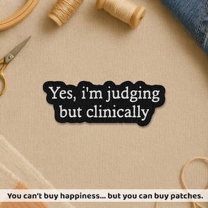 May include: Black patch with the text "Yes, I'm judging but clinically" in white. The patch is surrounded by sewing supplies, including thread spools, scissors, and a wooden embroidery hoop. The text "You can't buy happiness... but you can buy patches" is at the bottom.