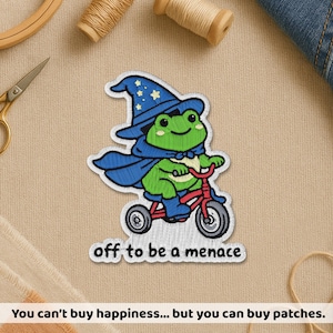 May include: Embroidered patch featuring a green frog wearing a blue wizard hat and cape, riding a red bicycle. The text "off to be a menace" is below the frog. The patch is on a beige fabric background.