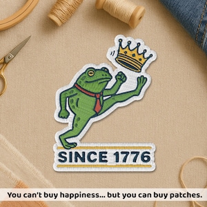 May include: Embroidered patch featuring a green frog wearing a red tie and a gold crown. The frog is leaping with one leg extended. The patch includes the text "SINCE 1776" and the phrase "You can't buy happiness... but you can buy patches."