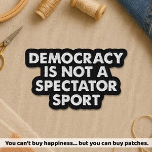 May include: Black and white embroidered patch with the text "DEMOCRACY IS NOT A SPECTATOR SPORT". The patch is rectangular with a curved bottom edge. Additional text at the bottom reads "You can't buy happiness... but you can buy patches."
