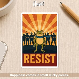 May include: A sticker featuring a cartoon frog wearing a blue scarf, standing in front of a group of police officers. The background has a sunburst design in orange and yellow, with the word "RESIST" in bold orange letters.