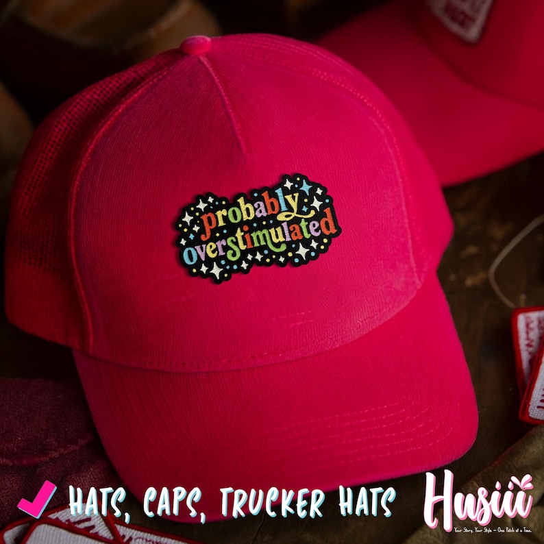 Op de afbeelding: Een felroze truckerpet met een mesh achterkant en een gebogen klep. De pet heeft een patch met de tekst "probably overstimulated" in kleurrijke letters. De afbeelding bevat ook de tekst "Hats, Caps, Trucker Hats".
