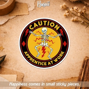 May include: A circular sticker with a yellow and black design. It features a skeleton with the text "CAUTION APPRENTICE AT WORK". The skeleton has blue eyes and is surrounded by red lightning bolts. The sticker has a white border.