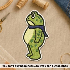 May include: Embroidered patch featuring a green frog with a blue scarf and a sad expression. The frog has green spots and a cream-colored belly. The text at the bottom reads: "You can't buy happiness... but you can buy patches."