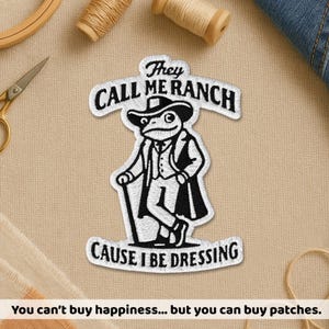 Peut inclure: Écusson brodé avec une grenouille portant un chapeau de cowboy et un costume, avec le texte "They Call Me Ranch Cause I Be Dressing." L'écusson est blanc avec des détails noirs. Le texte du bas dit "You can't buy happiness... but you can buy patches."