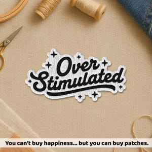 May include: Embroidered patch with the words "Over Stimulated" in black script on a white background. The patch has a black border and decorative crosses. The patch is on a beige surface with sewing supplies.