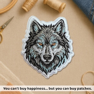 Peut inclure: Écusson brodé représentant un loup avec des détails de fourrure en nuances de gris, noir et sarcelle, avec des yeux orange. L'écusson a une bordure blanche et est posé sur un fond neutre. Le texte en bas dit : "On ne peut pas acheter le bonheur... mais on peut acheter des écussons."