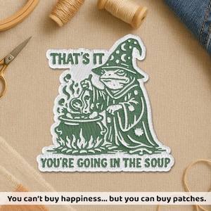 Peut inclure: Écusson brodé représentant un sorcier grenouille vert remuant un chaudron. Le texte "THAT'S IT" et "YOU'RE GOING IN THE SOUP" sont au-dessus et en dessous de la grenouille. L'écusson est blanc et vert.