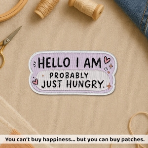 May include: A lavender and white embroidered patch with the text "HELLO I AM PROBABLY JUST HUNGRY." The patch has a decorative border with heart and star accents. The patch is on a beige fabric surface.