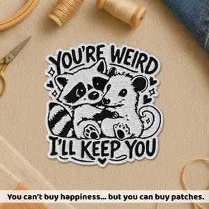 May include: Embroidered patch with a raccoon and possum hugging, with the text "YOU'RE WEIRD I'LL KEEP YOU" in black and white. The patch is surrounded by decorative elements, suitable for clothing or accessories.