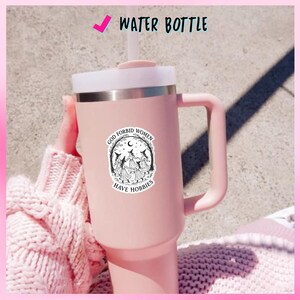 May include: A pink water bottle with a white lid and handle. The bottle has a sticker with the text "GOD FORBID WOMEN HAVE HOBBIES" and an illustration of witches. The words "WATER BOTTLE" are printed at the top.