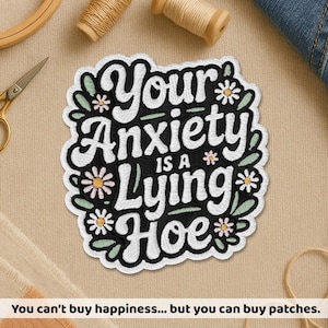 May include: Embroidered patch with the text "Your Anxiety is a Lying Hoe" in white script on a black background, surrounded by white and green floral accents. The patch is on a beige surface.