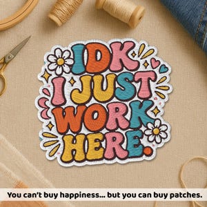 May include: Embroidered patch with the text "IDK I JUST WORK HERE" in colorful bubble letters. The patch has a white border with floral and heart accents. The bottom text reads "You can't buy happiness... but you can buy patches."
