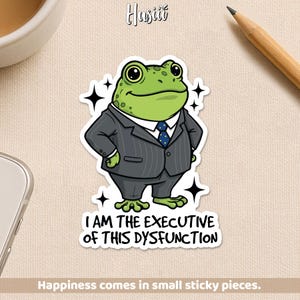 May include: A sticker featuring a cartoon frog in a gray pinstripe suit and a blue tie. The text reads "I AM THE EXECUTIVE OF THIS DYSFUNCTION." The sticker has a white border and star accents. The bottom text says "Happiness comes in small sticky pieces."