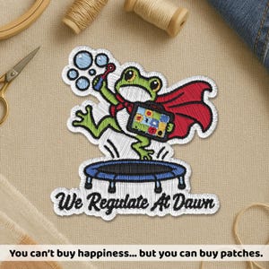 May include: An embroidered patch featuring a green frog wearing a red cape, jumping on a blue trampoline while holding a bubble wand and a colourful briefcase. The text "We Regulate At Dawn" is stitched in black below the frog on a white background.