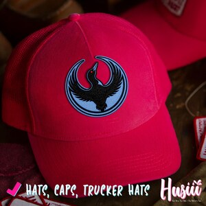 K&ouml;nnte beinhalten: Leuchtend pinkfarbene Trucker-Cap mit einem schwarzen Vogel-Emblem auf hellblauem Hintergrund. Der Vogel hat rote Augen und ist von einem hellblauen Ring umgeben. Die Cap hat einen Mesh-R&uuml;cken und einen gebogenen Schirm. Der Text auf dem Bild lautet "HATS, CAPS, TRUCKER HATS".