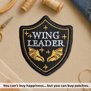 May include: Black shield-shaped patch with gold trim and the words "WING LEADER" in white. Features gold bat wings and cross accents. The text at the bottom reads "You can't buy happiness... but you can buy patches."