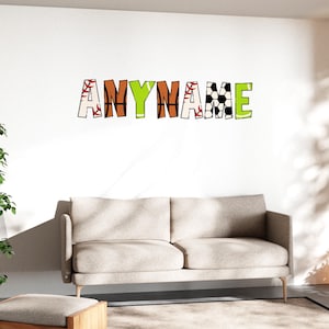 May include: A wall decal spelling "ANYNAME" in colorful letters. The letters are decorated with sports-themed patterns, including baseball stitching, basketball, soccer ball, and a green background. The decal is above a beige sofa.