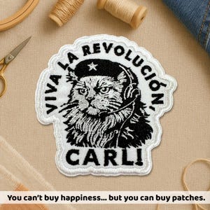 May include: Embroidered patch featuring a cat wearing a beret and headset, with the text "VIVA LA REVOLUCIÓN" and "CARLI" in black on a white background. The patch has a decorative border and is likely for clothing or accessories.