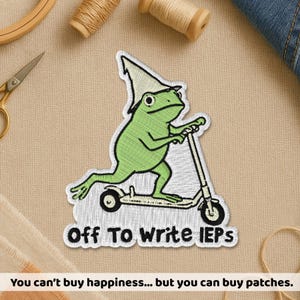 May include: Embroidered patch featuring a green frog riding a scooter, wearing a pointed hat. The text "Off To Write IEPs" is below the frog. The bottom of the patch reads "You can't buy happiness... but you can buy patches."