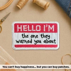 May include: Rectangular embroidered patch with the text "HELLO I'M the one they warned you about" in white on a red and white background. The patch has a white border. Sewing supplies are in the background.